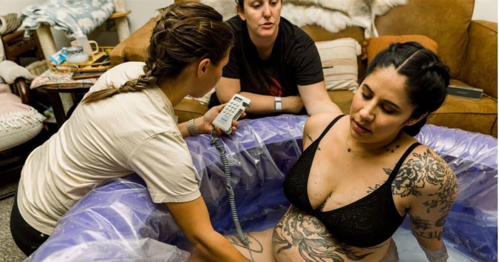 Birthing person laboring in water at home with midwive checking heart tones Photo by Luna Birth https://www.lalunabirth.com/
