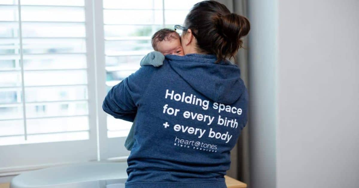 Winner of the Best Doula Award Salt Lake City 2024 wearing the Heart Tones Birth motto hoodie & holding a newborn baby