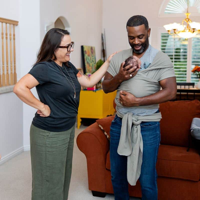Postpartum Doula Beth Hardy supports new dad at home holding baby in sling in Salt Lake City
